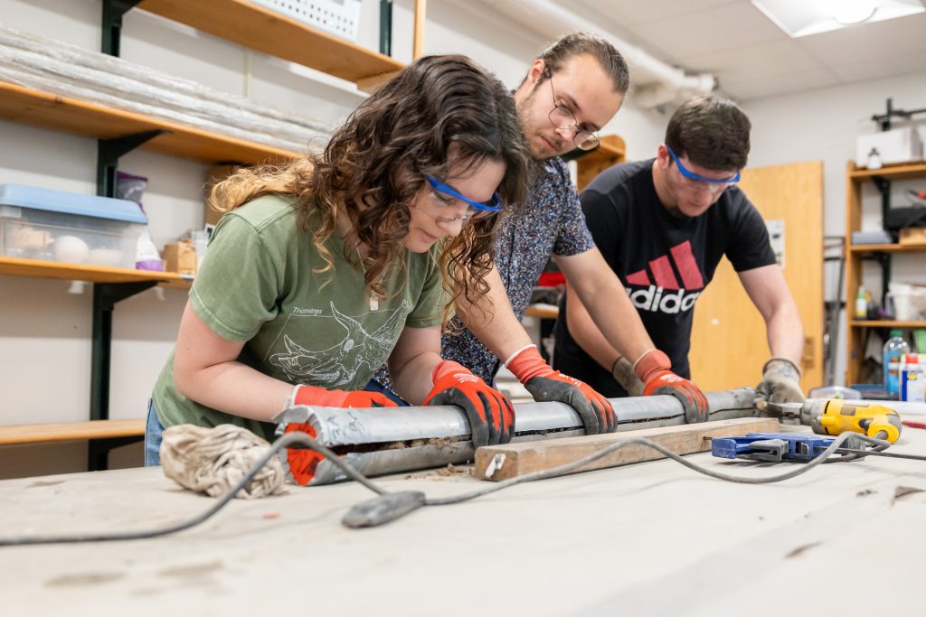 From left, Cassie Aimetti '25 (CLAS), Preston Senderoff '25 (CLAS) '22 (CLAS), and Zak Knowlton '25 (CAHNR) split a soil core their team collected on Grannis Island in New Haven in one of the soil core labs in Beach Hall on June 6, 2024. (Sydney Herdle/UConn Photo)