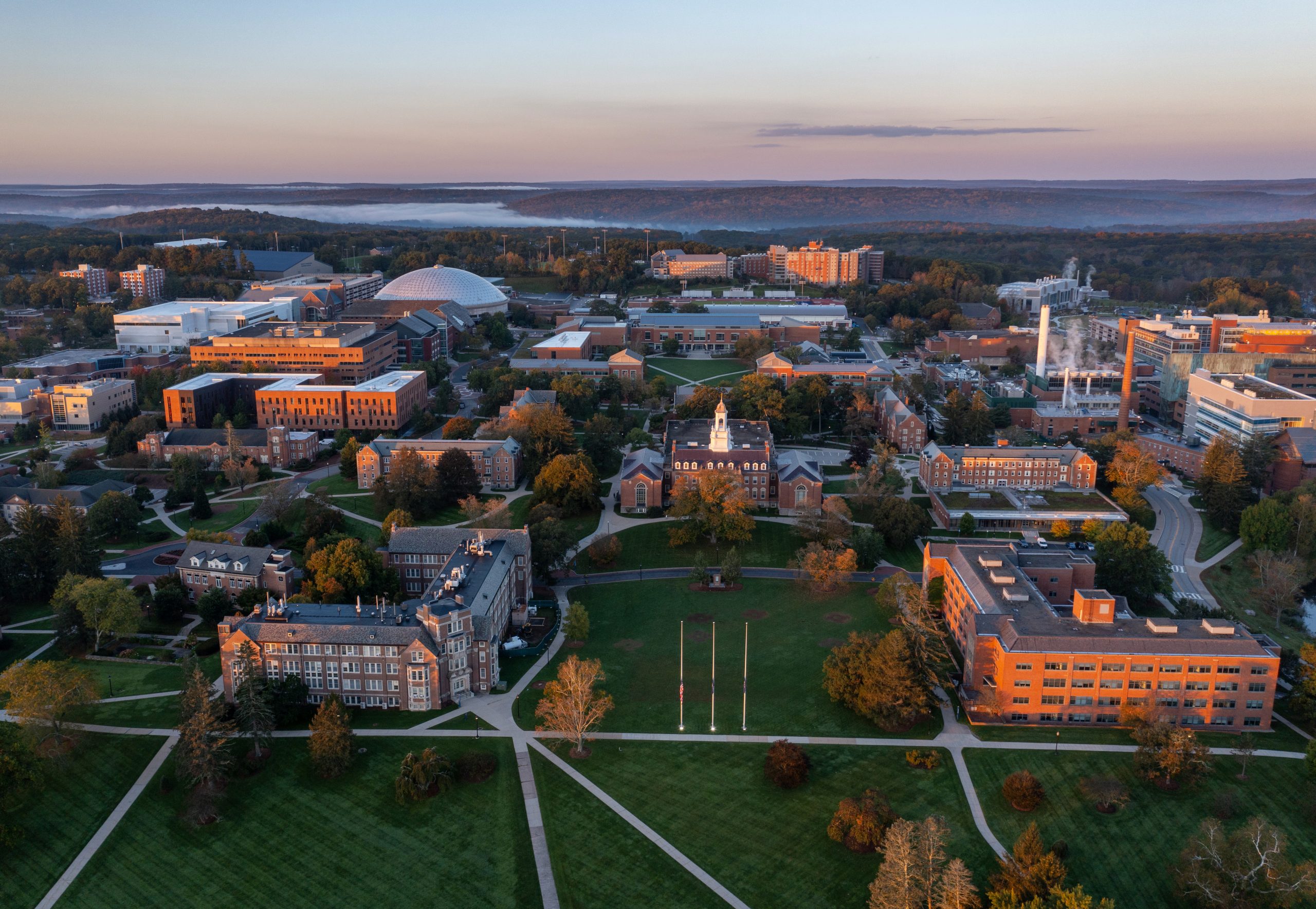 Aerial view of the UConn Storrs campus, showing campus buildings, roads, green spaces, and the surrounding landscape.